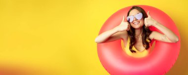 Close up portrait of happy attractive woman in sunglasses, wearing pink swimming ring, smiling and showing thumbs up, standing over yellow background.