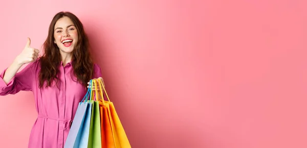 年轻快乐的女购物者竖起大拇指 对打折很满意 买了便宜货 拿着购物袋 高兴地微笑着 粉红的背景 — 图库照片