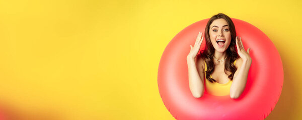 Beautiful coquettish woman inside pink swimming ring, posing on summer vacation, beach holiday concept, standing against yellow background.
