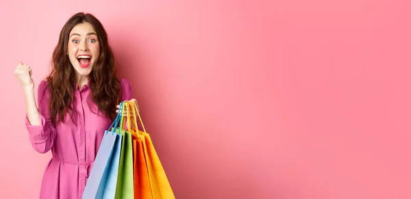 興奮した女の子は喜びの叫び 拳ポンプを作り ショッピングバッグを保持し ピンクの背景の上にドレスに立って — ストック写真