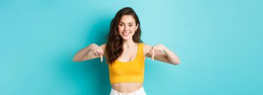 Yönünü gösteren güzel bir genç kadının portresi, parmaklarıyla reklamı işaret ediyor, neşeli bir şekilde gülümsüyor, çıkış öneriyor, mavi arka planda duruyor..