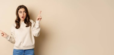 Confused girl shopper pointing sideways, making choice, decision between two variants, beige background. Copy space