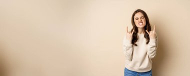 Happy young woman enjoying music, having fun, showing rock on, heavy metal finger horns gesture, standing over beige background.