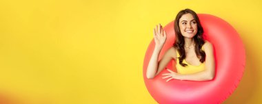 Happy young woman wearing pink swimming ring, waving hand and saying hi, enjoying vacation, summer beach holiday, standing over yellow background.