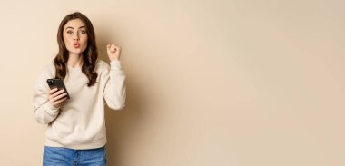 Happy girl holding smartphone and celebrating, winning on mobile phone app, standing over beige background. Copy space