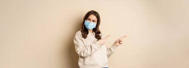 Health and covid-19 concept. Confused young woman in medical face mask, pointing left and staring with frustrated face, beige background.