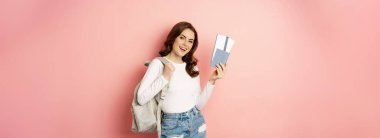 Vacation and holiday. Beautiful girl going on trip, holding passport with airplane tickets, holding backpack, travelling, standing over pink background.