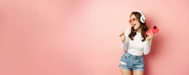 Summer girl laughing, eathing lolipop and listening music in headphones, dancing with smartphone against pink background.