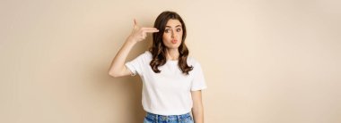 Young woman showing finger pistol sign near head and looking annoyed, sick and tired, standing over beige background.