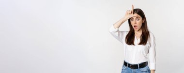 Young woman mocking loser, showing L word sign on forehead and gasping with disbelief surprise, standing against white background.