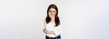 Beautiful young brunette woman, gently touches her face skin and smiling coquettish, tender pose against studio white background.