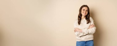 Beautiful young brunette woman posing happy against beige background, smiling at camera, wearing sweater.