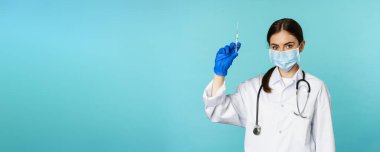 Covid ve sağlık hizmetleri konseptinden aşı. Genç kadın doktor, maske ve eldivenli hemşire aşı yapmak için şırınga kullanıyor, torkuaz arka planda duruyor..