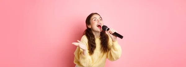 Happy young woman enjoying playing karaoke, singing in mic, looking aside at screen with lyrics, smiling cheerful, standing against pink background.