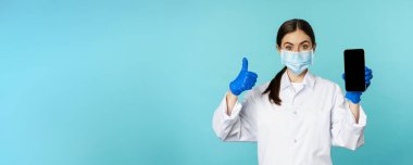 Portrait of doctor in medical face mask and gloves, showing mobile phone app, smartphone screen and thumb up, recommending online checkup website, standing over blue background.