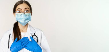 Şaşırmış ve şaşırmış genç bir doktorun portresi, hayranlıkla bakıyor, kalbe el ele tutuşuyor, beyaz arka planda tıbbi yüz maskesi takıyor..