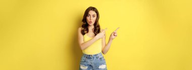 Portrait of stylish summer girl, brunette woman showing way, pointing fingers left, advertising, standing over yellow background. Copy space