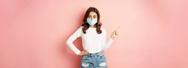 Beautiful stylish woman in medical face mask from covid, pointing finger at upper left corner, showing advertisement, standing over pink background.