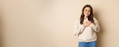 Smiling woman holding hands on heart, looking with care and love at camera, standing in sweater over beige background.