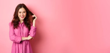 Güzel esmer kadın bahardan zevk alıyor, parlak giysiler giyiyor, kıvırcık saçlarına dokunuyor ve kameraya gülümsüyor, pembe arka planda duruyor..