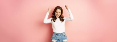 She is a winner. Enthusiastic brunette girl celebrating, jumping from happiness and shouting joyful, achieve goal success, standing over pink background. Victory concept