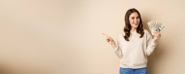 Smiling beautiful woman holding money dollars and pointing finger left, showing logo company banner, standing over beige background.
