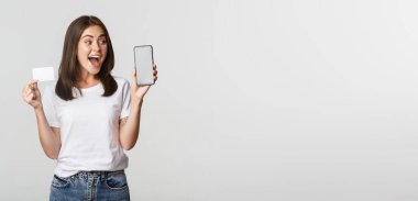 Heyecanlı ve şaşırmış güzel kız ekranda kredi kartı ve cep telefonu bankacılık uygulaması gösteriyor..