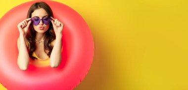 Close up portrait of stylish young woman, girl with swimming ring and summer sunglasses, ready for beach holiday, vacation, standing over yellow background.