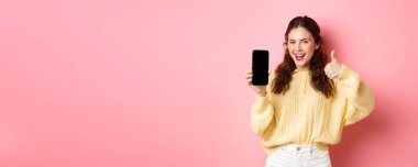 Çok iyi bir uygulama. Gülümseyen tatmin olmuş kadın başparmağını kaldırıyor ve akıllı telefon ekranını boşaltıyor, uygulama veya web sitesi öneriyor, pembe arka plana karşı duruyor.