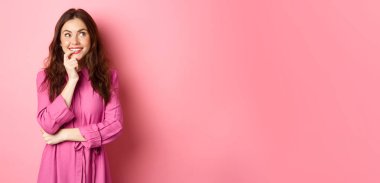 Düşünceli, düşünceli genç bir kadın. Gülümsüyor ve parmağını ısırıyor. Düşünürken, hayal ederken, pembe arka planda dikilirken..