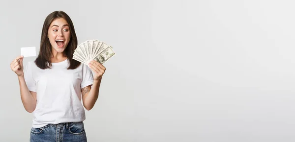 Rozbawiona Piękna Dziewczyna Patrząc Pieniądze Trzymając Kartę Kredytową Stojące Białe — Zdjęcie stockowe