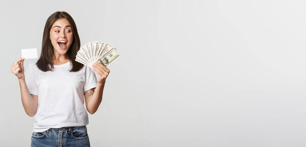 Amused beautiful girl looking at money and holding credit card, standing white background.