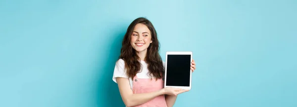 Image of happy young girl showing digital tablet screen and smiling proud, showing your logo on display, standing over blue background.