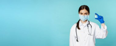 Aşı kampanyası. Yüzünde tıbbi maske ve eldiven olan gülümseyen kadın doktor Covid, Omicron delta varyantı aşısı gösteriyor. Torkuaz arka planda duruyor..