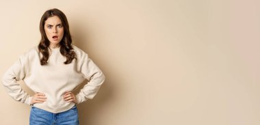 Frustrated, offended young woman looking hurt ad insulted at camera, standing shocked over beige background. People and emotions concept