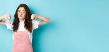 Şüpheci genç kızın portresi hayır diyor, baş parmaklarını aşağı gösteriyor, kötü reklam anlaşmasından hoşlanmıyor, olumsuz cevap veriyor, mavi arka planda duruyor..