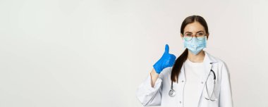 Coronavirus 'un yüz maskesi takmış kadın doktorların portresi. Baş parmaklarını gösterip gülümsüyor. Klinik ya da tıbbi web sitesi tavsiye ediyor..