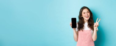 Sevimli genç bir kadın gülümsüyor ve boş akıllı telefon ekranı ile V işareti gösteriyor, uygulamayı veya mobil mağazayı gösteriyor, mavi arka planda duruyor..
