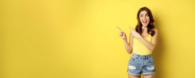 Portrait of stylish summer girl, brunette woman showing way, pointing fingers left, advertising, standing over yellow background. Copy space