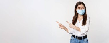Tıbbi maskeli kurumsal bir kadının portresi, solu işaret ediyor, logoyu, şirket afişini gösteriyor, beyaz arka planda duruyor..