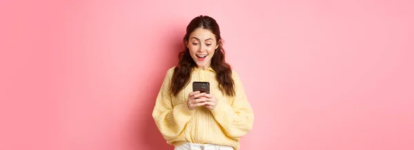 兴奋的黑发女人惊讶地喘息 在智能手机上看到信息 看着手机 站在粉色背景上的画像 — 图库照片