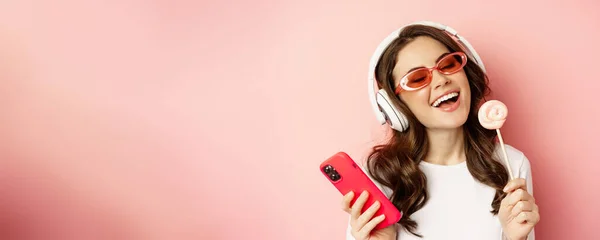 Beautiful female model listening music in headphones, holding lolipop and mobile phone, posing in sunglasses, standing over pink background.