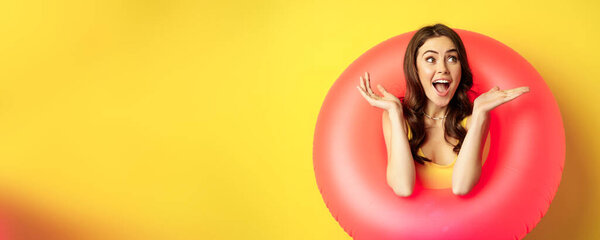Happy beautiful young woman relaxing on summer vacation, wearing pink swim ring for beach holiday, standing against yellow background.