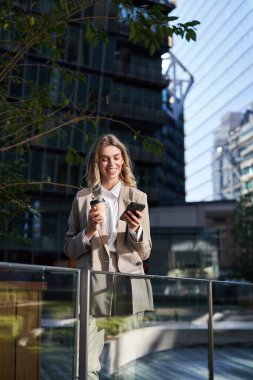 Takım elbiseli bir kadın, kahve içiyor, cep telefonunu elinde tutuyor, tek başına şehirde duruyor..