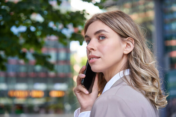 Close up shot of businesswoman talking on mobile phone. Corporate woman calling someone, looking around, standing outdoors.