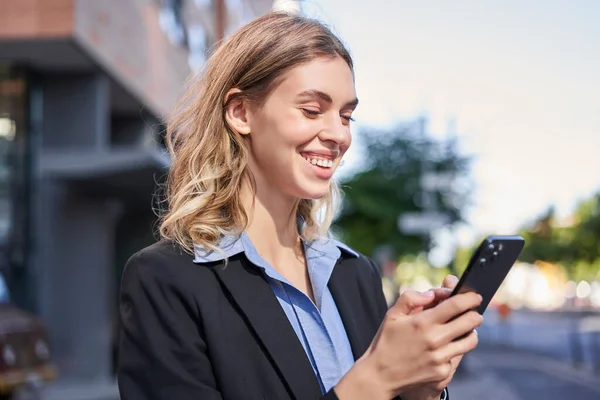 Corporate woman stands on street and texts message on mobile phone, smiling while looking at smartphone screen.