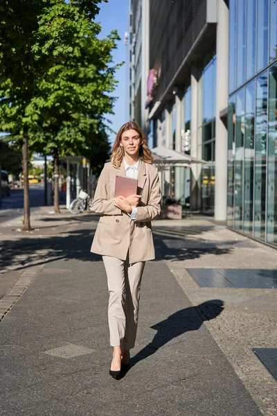 Vertical shot of businesswoman walking on street with digital tablet, going to work, wearing beige suit and high heels.