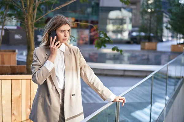 Businesswoman with concerned face, talks on mobile phone and stands on street, calling business partner.