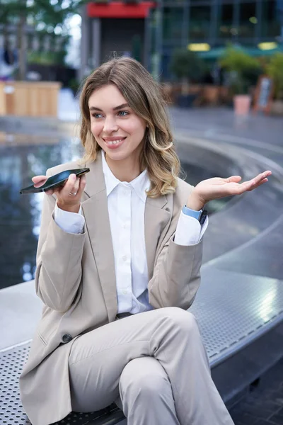 Smiling business woman record voice message, speaking into microphone on mobile phone, sitting near fountain on street.