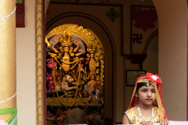 Howrah, Hindistan -26 Ekim 2020: Bengalli kız çocuğu şenlikli elbiseler içinde, arka planda Tanrıça Durga ile gülümsüyor ve poz veriyor, yaşlılığın içinde dekore edilmiş ev. Durga Puja, en büyük Hinduizm festivali..